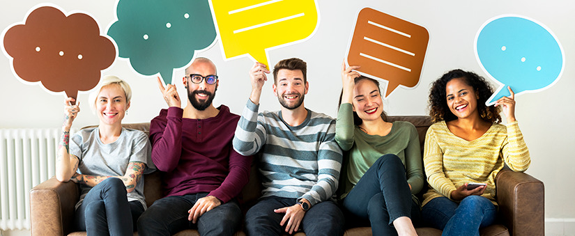 Cinco pessoas com roupas coloridas sentadas num sofá marrom seguram plaquinhas de balões de palavra para cima.