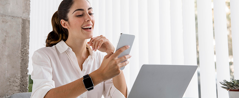 Atendimento ao cliente Mulher olhando para um celular sorrindo e um notebook à sua frente.