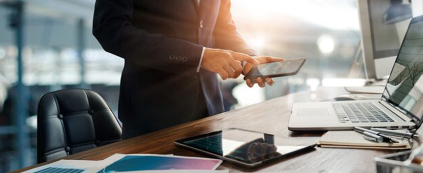 gestão de vendas lançamento imobiliário: imagem de uma mesa com alguns dispositivos digitais e um homem ao lodo com o celular na mão.