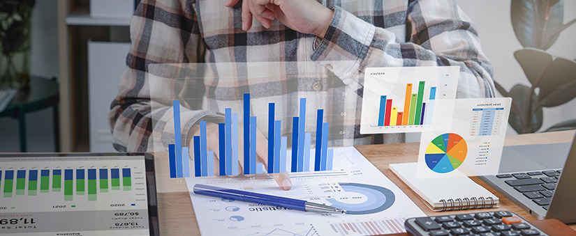 reduzir custos com marketing digital reduzir custos: imagem de uma mesa com alguns papeis e dispositivos digitais, e alguns hologramas com gráficos. Ao fundo uma pessoa aponta para um dos papeis.