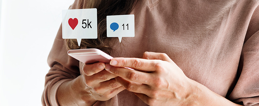 influência das mídias sociais close nas mãos de uma pessoa segurando um celular. Acima do celular ícones de notificação de like e de chat.