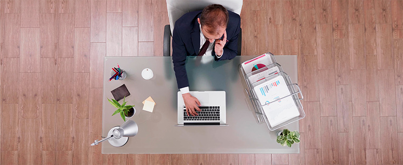 gestão eficiente para o mercado imobiliário imagem de cima de um homem de terno sentado à frente de uma mesa com um notebook, papeis com gráficos e itens de escritório.