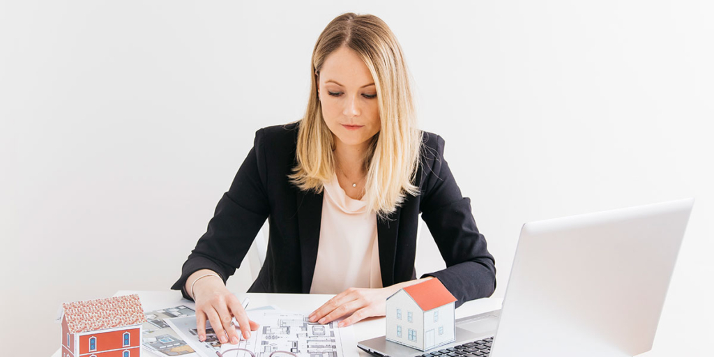 Site de imóveis Site de imóveis: imagem de uma mulher sentada à frente de uma mesa com um notebook, alguns papeis e miniatura de casas de papel.