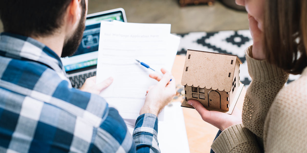 vistoria de apartamento novo vistoria de apartamento novo: um homem e uma mulher sentados, de costas, olhando para alguns papeis. Na mão da mulher uma casa de madeira em miniatura.