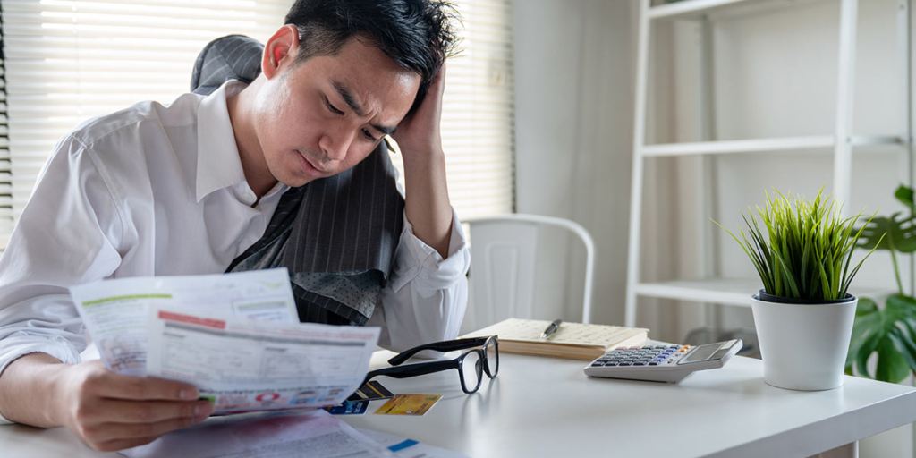 adimplência adimplência: homem sentado à frente de uma mesa com alguns papeis numa das mãos e aparentemente preocupado.
