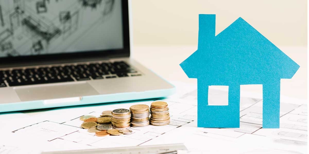 investir no mercado imobiliário investir no mercado imobiliário: imagem de uma casa de papel ao lado de algumas moedas empilhadas. No fundo um notebook mostrando a planta de uma casa.