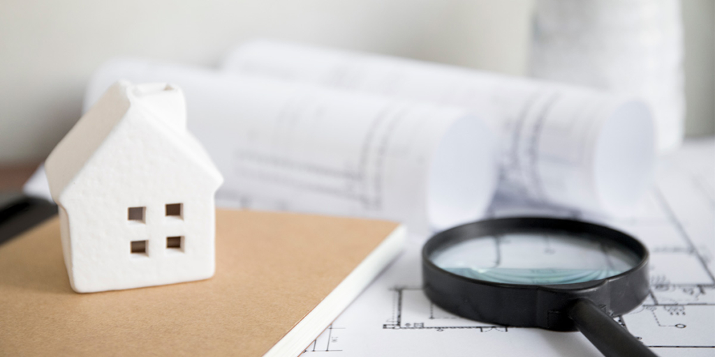 Norma abnt para avaliação de imóveis: imagem de uma mesa com uma planta de imóvel em papel, acima uma lupa e uma miniatura de casa.