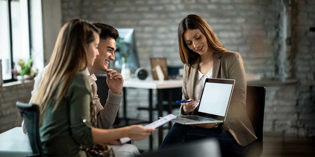 O que é permuta de imóveis: imagem de uma mulher segurando um notebook e apontando para a tela, mostrando algo para mais duas pessoas à sua frente.