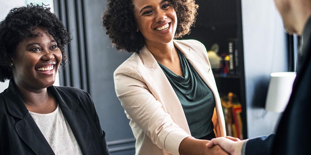 Onboarding de clientes: Duas mulheres de terno sorrindo para uma pessoa de costas enquanto uma delas aperta sua mão.