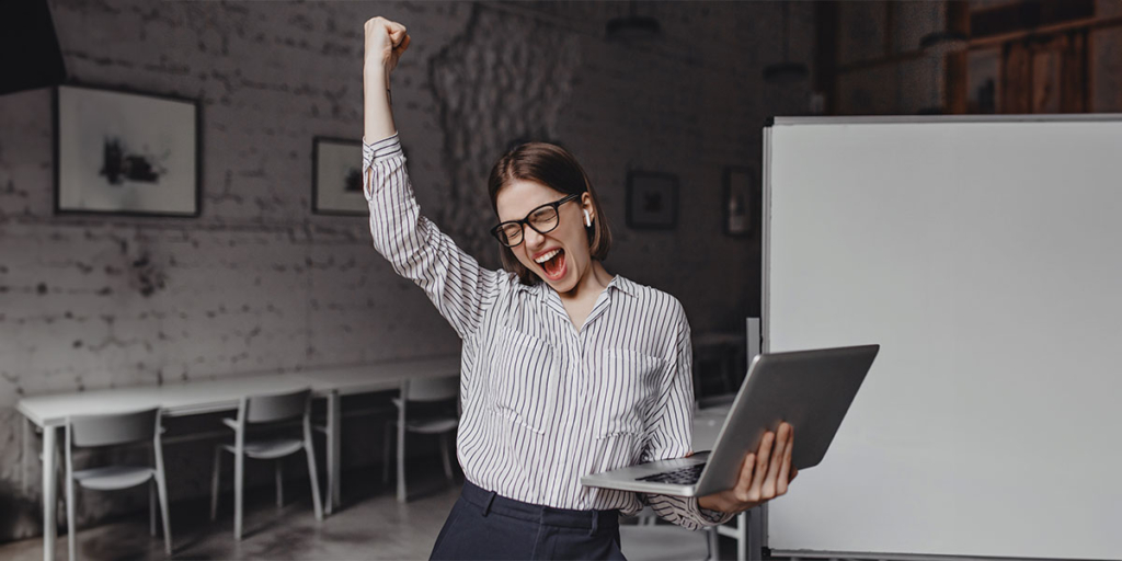 Script para corretor de imóveis Script para corretor de imóveis: Imagem de uma mulher segurando um notebook com um dos braços levantados em comemoração.