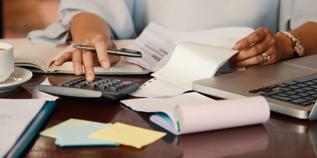 Contas a receber: close nas mãos de uma pessoa na frente de uma mesa com vários papeis, uma calculadora, notebook e uma xícara de café.