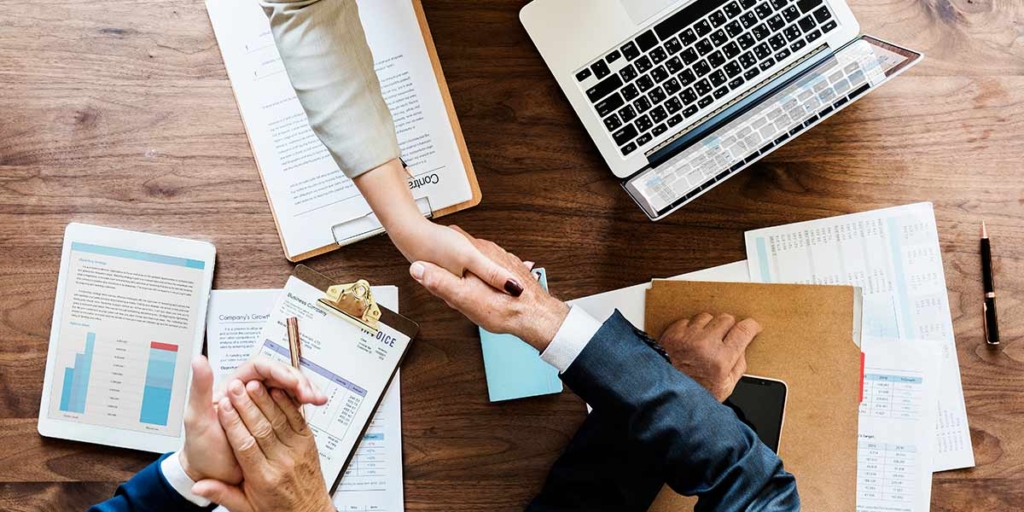 gestão de contratos gestão de contratos: imagem de uma mesa vista de cima com alguns papeis, um notebook e, no centro, foco num aperto de mão entre duas pessoas.