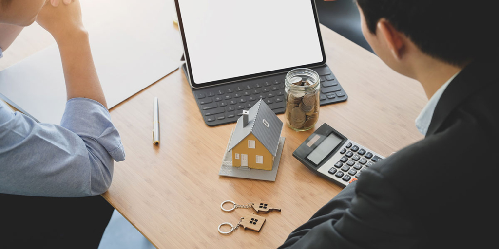 Captador de imóveis Captador de imóveis: imagem de uma mesa vista de cima e duas pessoas com os braços apoiados. Na mesa um tablet, uma calculadora, uma miniatura de casa e algumas moedas num pote de vidro.