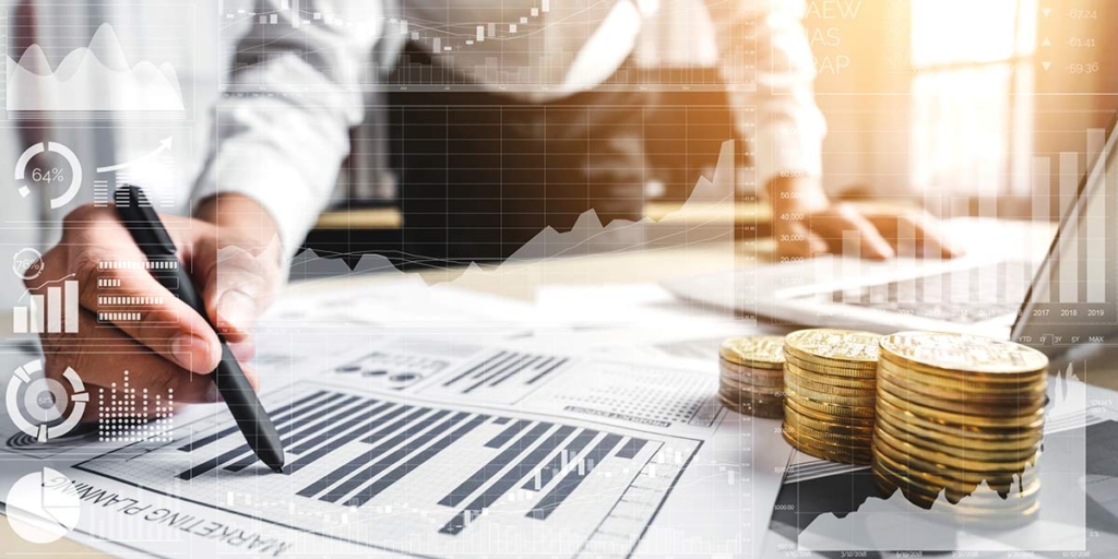 O que é lig investimento O que é lig investimento: close nas mãos de uma pessoa em cima de uma mesa cheia de papeis, moedas empilhadas e um notebook.