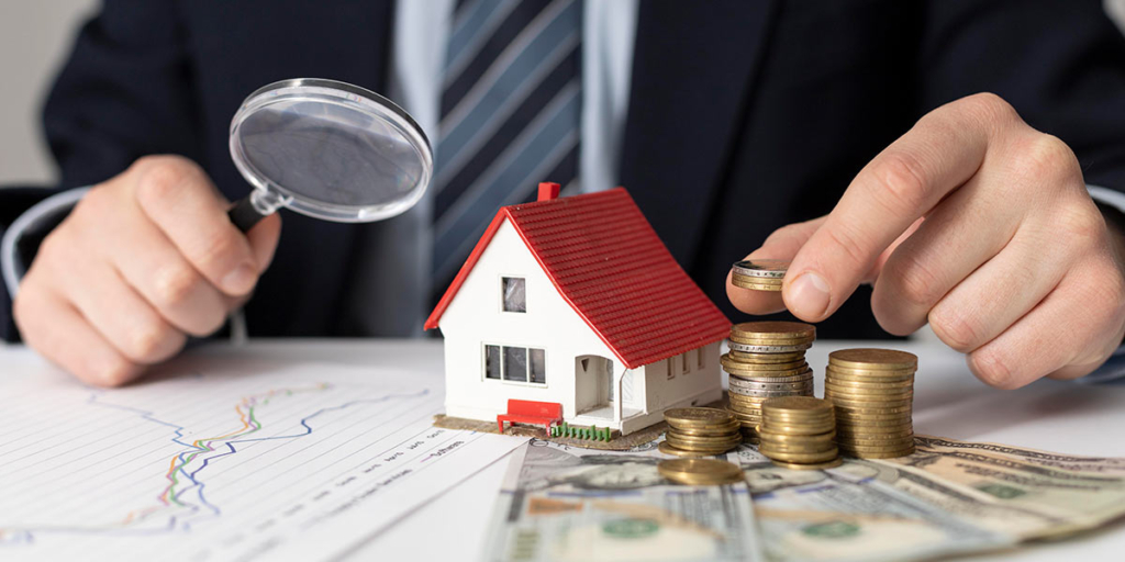 O que é lig investimento: close nas mãos de uma pessoa de terno em cima de uma mesa segurando uma lupa. Na mesa a miniatura de uma casa e algumas moedas empilhadas.