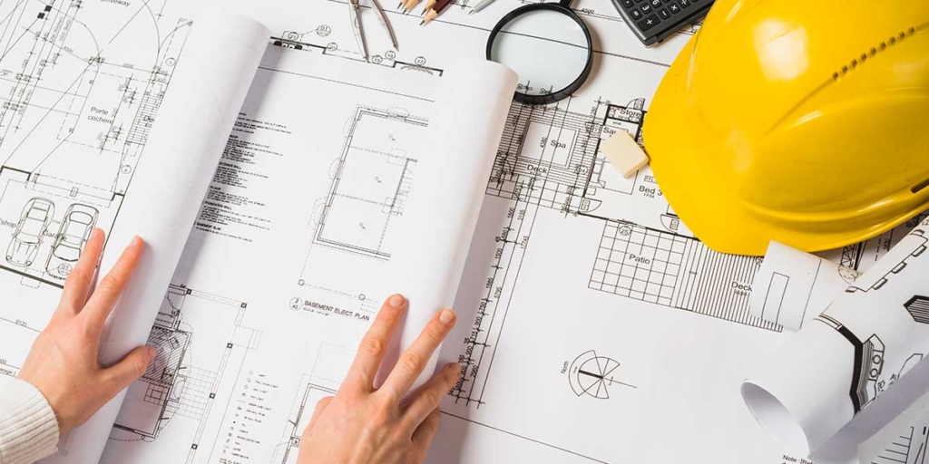 Construtoras no Brasil: mãos abrindo um projeto de planta baixa. Ao lado, aparece um capacete de construção amarelo