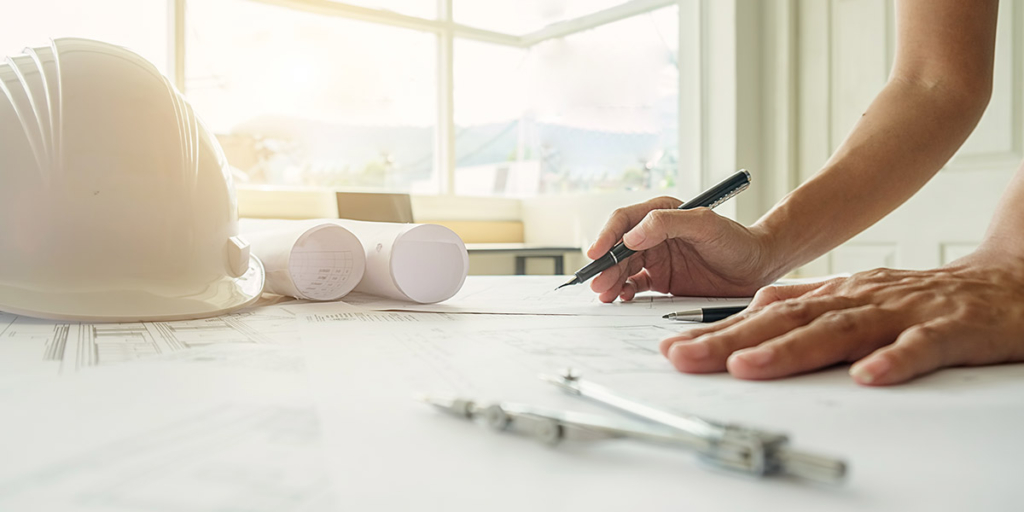 Como uma incorporadora ganha dinheiro Como uma incorporadora ganha dinheiro: close nas mãos de uma pessoa em cima de uma mesa segurando um lápis em cima de vários papeis. Na mesa também um capacete de proteção.