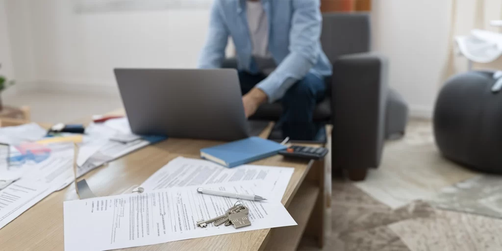 Documento de venda de imóvel Documento de venda de imóvel: imagem de uma mesa com muitos papeis e uma chave em destaque. Ao fundo uma pessoa num notebook.