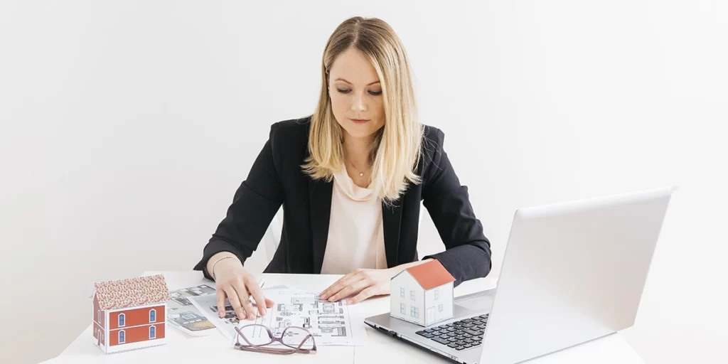 Gestor Imobiliário Gestor Imobiliário: imagem de uma mulher sentada em frente a uma mesa com muitos papeis, um óculos de grau, miniaturas de casa e um notebook.