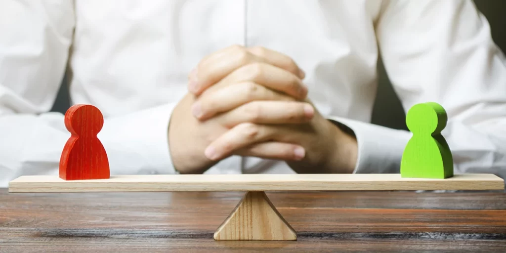 Mediação Imobiliária: imagem de uma gangorra de madeira com bonecos de madeira de cores diferentes de cada lado. Ao fundo vemos as mãos de uma pessoa entrelaçadas.