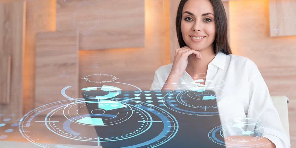 O que é um assistente virtual O que é um assistente virtual: imagem de uma mulher em frente a um notebook olhando para a câmera e sorrindo.