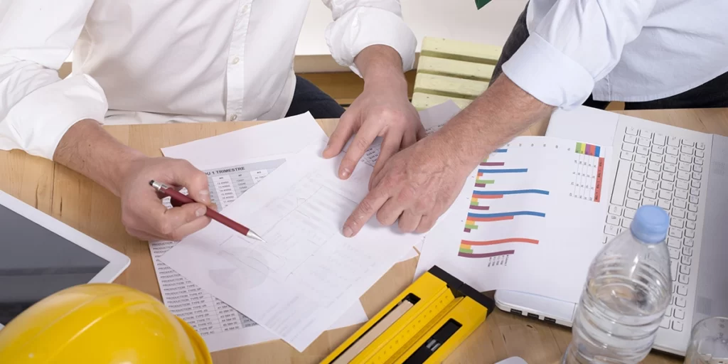 Tipos de INCC Tipos de INCC: imagem de uma mesa com muitos papeis com gráficos, um notebook e alguns itens de construção civil como um capacete e uma régua. Em destaque, mãos de duas pessoas apontando para os papeis.