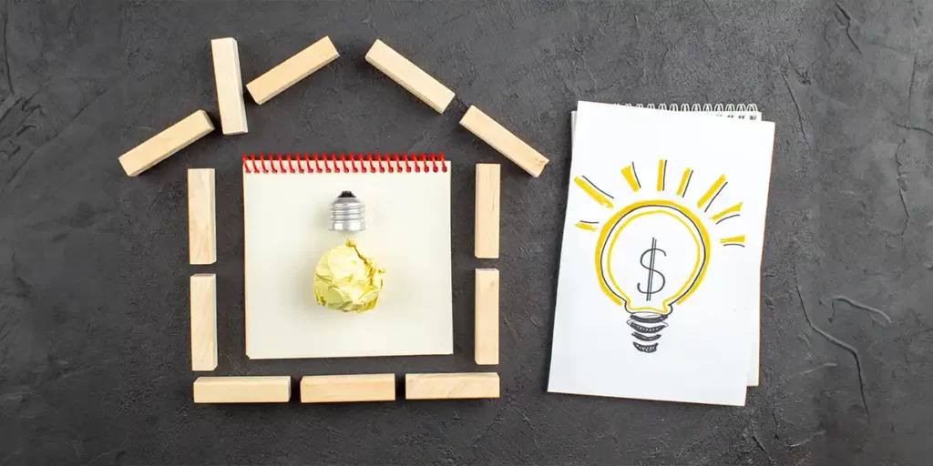 Captação de clientes no mercado imobiliário Captação de clientes no mercado imobiliário: do lado esquerdo imagem de blocos de madeira numa mesa formando uma casa. No centro, um caderno com a representação de uma lâmpada. Do lado direito um caderno aberto com a ilustração feita à mão de uma lâmpada acesa com um $ no centro.