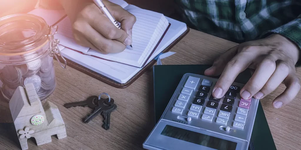 Carteira de corretor de imóveis: imagem de uma mesa vista de cima. Vemos as mãos de alguém manuseando uma calculadora e, com a outra mão, escrevendo num papel. Na mesa vemos um pote com muitas moedas dentro.