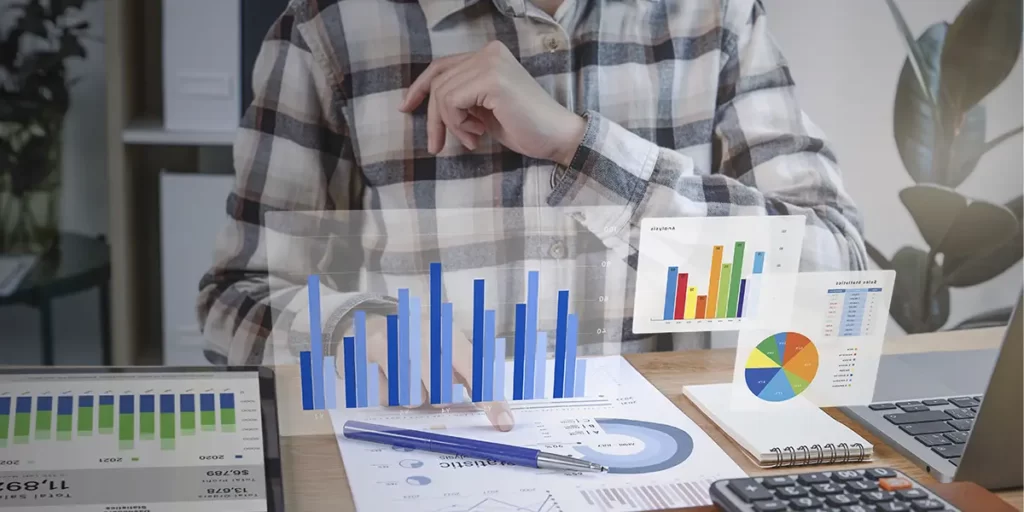 Indicadores de satisfação do cliente Indicadores de satisfação do cliente: close nas mãos de uma pessoa sobre uma mesa com muitos papeis, calculadora, um notebook. Acima, em destaque, ilustrações de gráficos.