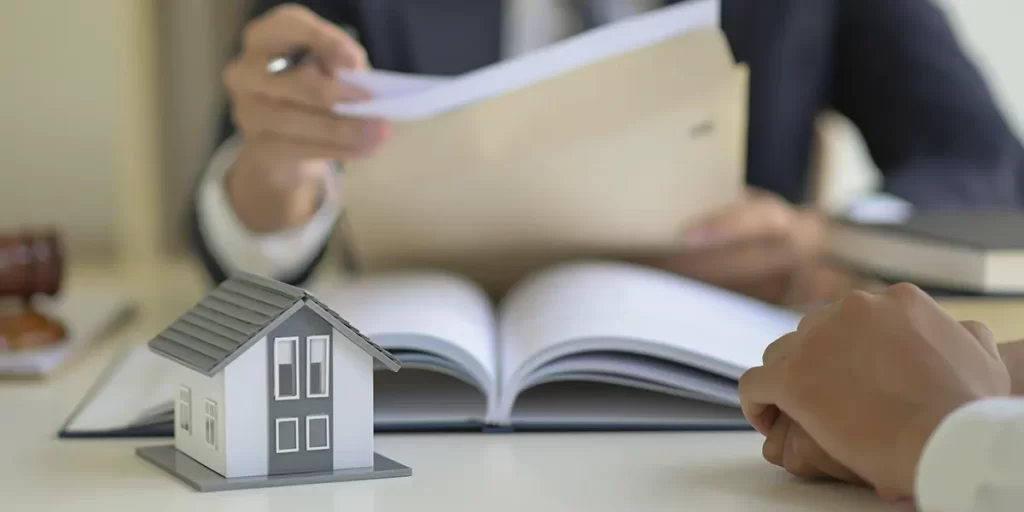 O que é averbação de um imóvel: imagem das mãos de duas pessoas numa mesa. No centro alguns papeis e, em foco, a miniatura de uma casa.