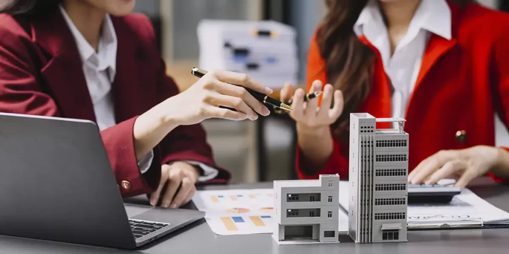 PropCo PropCo: foco nas mãos de duas mulheres gesticulando, com lápis na mão. Elas estão num escritório, na frente de uma mesa com papeis com gráficos, notebook e miniatura de edifícios.