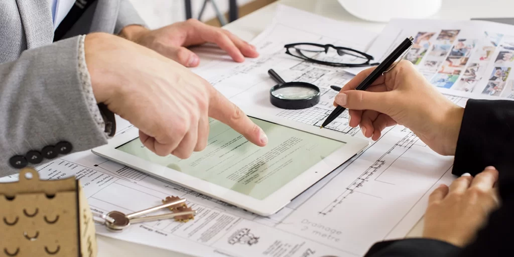 Vendas Consultivas Vendas Consultivas: close nas mãos de duas pessoas numa mesa com muitos papeis. Uma delas aponta para um tablet.