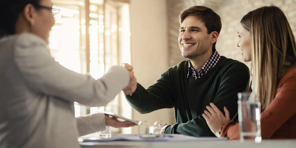 conexão com o cliente conexão com o cliente: imagem de um casal (homem e mulher) de um lado de uma mesa, sorridentes. Um deles aperta a mão de uma mulher do outro lado da mesa.