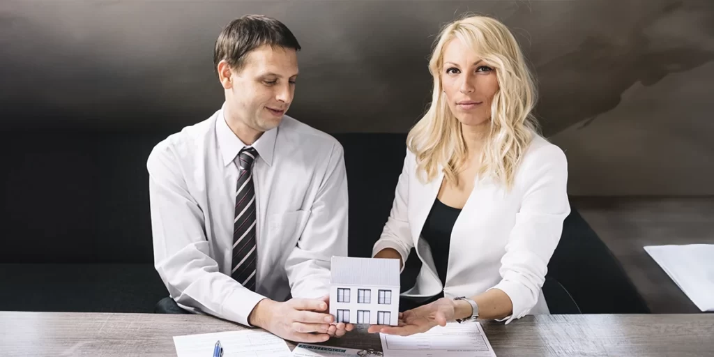 Código de Ética do corretor de imóveis Código de Ética do corretor de imóveis: imagem de duas pessoas diante de uma mesa. As duas usam terno e seguram a miniatura de uma casa.