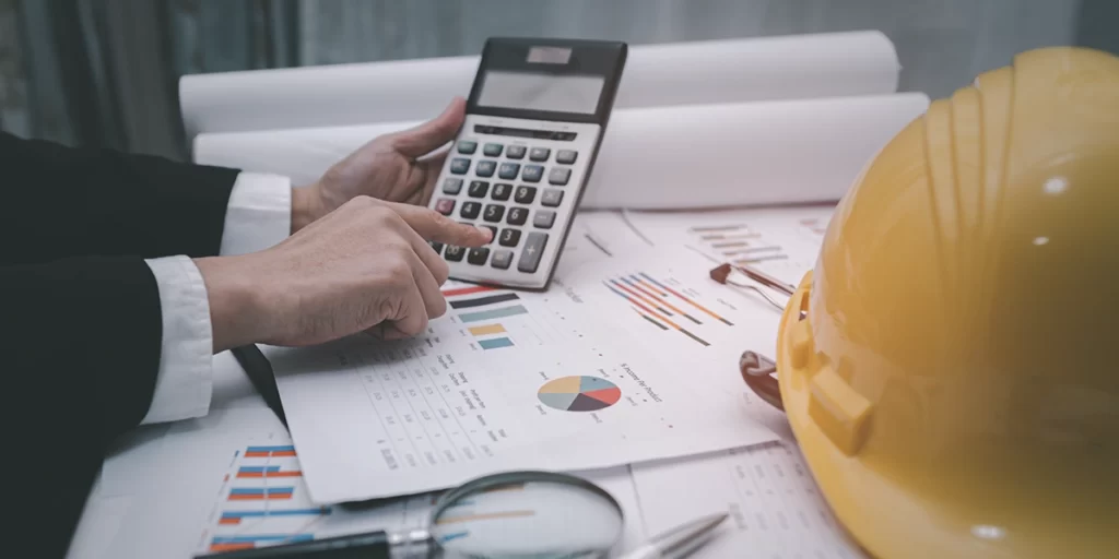 Como calcular custo de obra Como calcular custo de obra: close nas mãos de uma pessoa segurando uma calculadora. As mãos estão apoiadas na mesa com muitos papeis com coloridos gráficos. Na mesa também vemos um capacete de proteção em canteiro de obras.