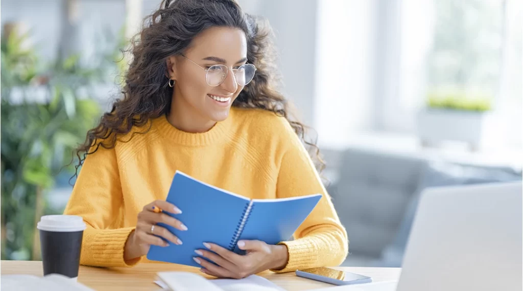 Cursos para corretor de imóveis Cursos para corretor de imóveis: imagem de uma mulher sorridente, sentada à frente de uma mesa. Ela segura um caderno azul e olha para um notebook.