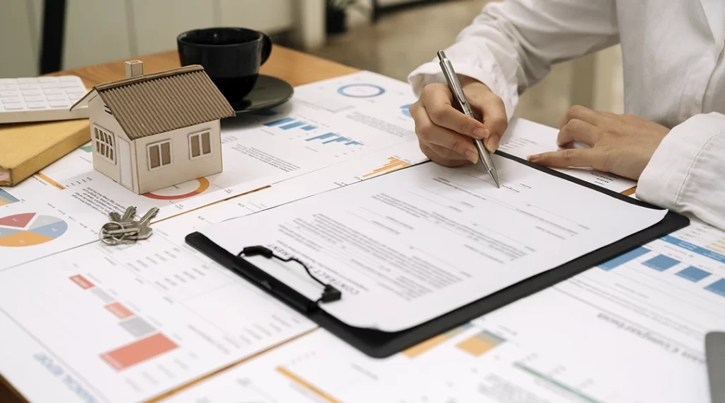 Cursos para corretor de imóveis Cursos para corretor de imóveis: close nas mãos de uma pessoa assinando um papel numa mesa com muitos papeis com gráficos espalhados. De um dos lados uma casa em miniatura e uma chave.
