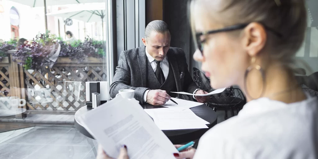 Ficha de visita de Imóvel Ficha de visita de Imóvel: imagem de um ambiente de café. Numa mesa um homem, que veste um terno, está olhando alguns papeis. Em primeiro plano, em desfoque, uma mulher também olha um papel.