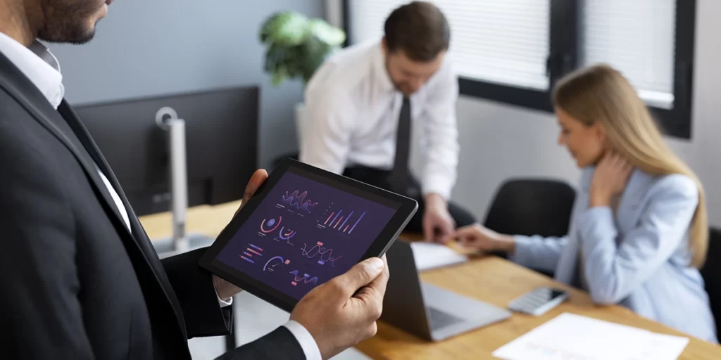 Portifólios imobiliários Portifólios imobiliários: imagem de um homem num escritório segurando um tablet com um dashboard na tela. Em segundo plano um homem e uma mulher conversam.