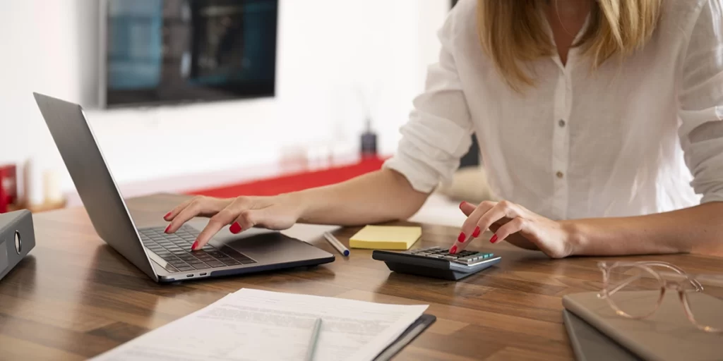 RRT RRT: close nas mãos de uma mulher, que está diante a uma mesa. Com uma das mãos ela usa uma calculadora, com a outra digita num notebook.