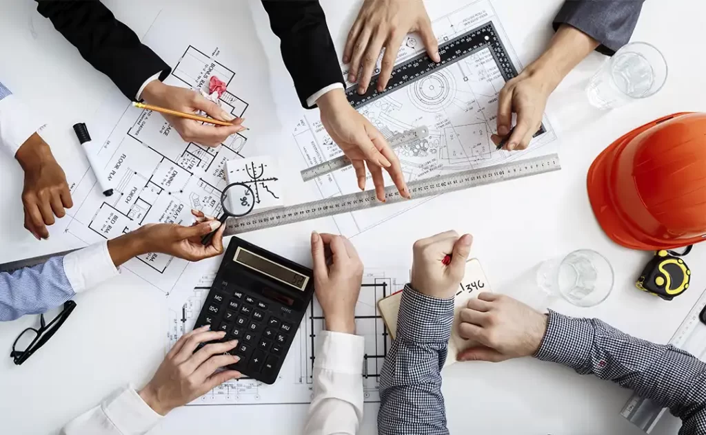 Administração de Construtora Administração de Construtora: imagem vista de cima, de uma mesa com muitos papeis e mãos de várias pessoas segurando réguas, calculadoras, canetas e plantas de imóveis.