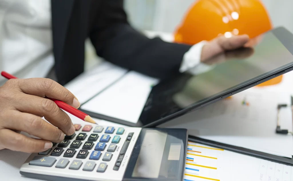 Administração de Construtora Administração de Construtora: close nas mãos de uma pessoa em cima de uma mesa com muitos papeis. Com uma das mãos ela segura uma calculadora e, na outra um tablet.