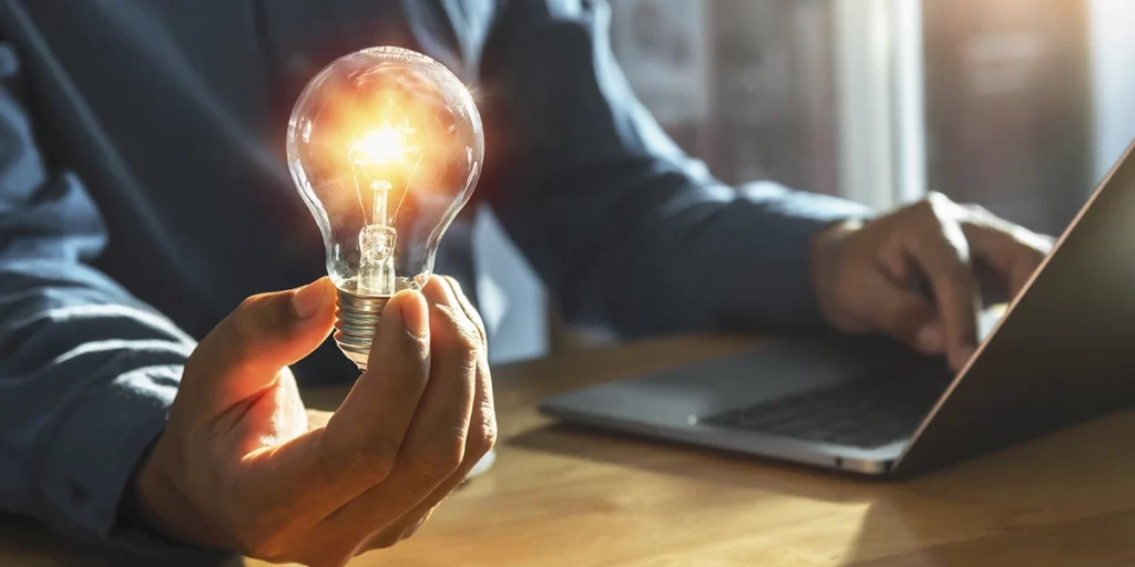 Anúncio Imobiliário Anúncio Imobiliário: close nas mãos de uma pessoa usando terno, sentada diante de uma mesa, digitando no teclado de um notebook. Com a outra mão, ela segura uma lâmpada acesa.