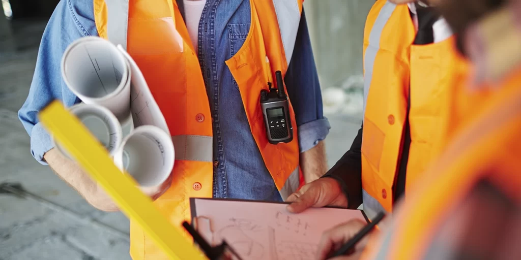 Gestão de Resíduos na Construção Civil Gestão de Resíduos na Construção Civil: close nas mãos de duas pessoas segurando plantas de imóveis, pranchetas, régia e lápis, usando coletes laranjas.