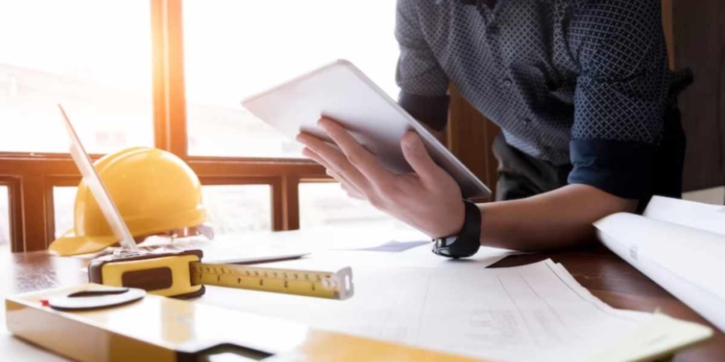 Mobilização e Desmobilização Mobilização e Desmobilização: close na mão de uma pessoa segurando um tablet diante de uma mesa com muitos papeis, um capacete de proteção e uma régua.