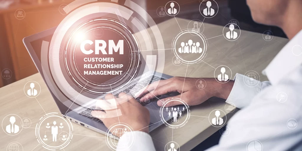 Software Imobiliário Software Imobiliário: close nas mãos de uma pessoa digitando num notebook. Em destaque na imagem, a sigla "CRM" e algumas ilustrações de pessoas em volta.