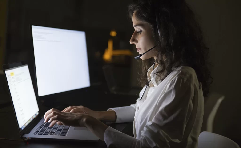 vantagens da inteligência artificial vantagens da inteligência artificial: imagem de uma mulher sentada à frente de um computador numa sala com poucas luzes, destacando as luzes das telas na mesa. Ela sua um fone com microfone.