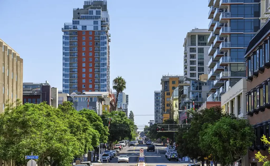 Indicadores imobiliários Indicadores imobiliários: A imagem mostra uma vista de uma rua em um ambiente urbano. Há edifícios altos ao longo da borda da rua, incluindo prédios residenciais e possivelmente comerciais. A rua está bem iluminada e parece ser um dia claro. Há muitos carros estacionados e em movimento, e a paisagem é acentrada pela presença de árvores ao longo da borda da rua, o que dá um contraste verde à cena urbana. A rua é ampla e parece ser uma via principal, com sinais de trânsito e possivelmente uma estrutura de transporte aéreo ou ferroviário ao fundo.