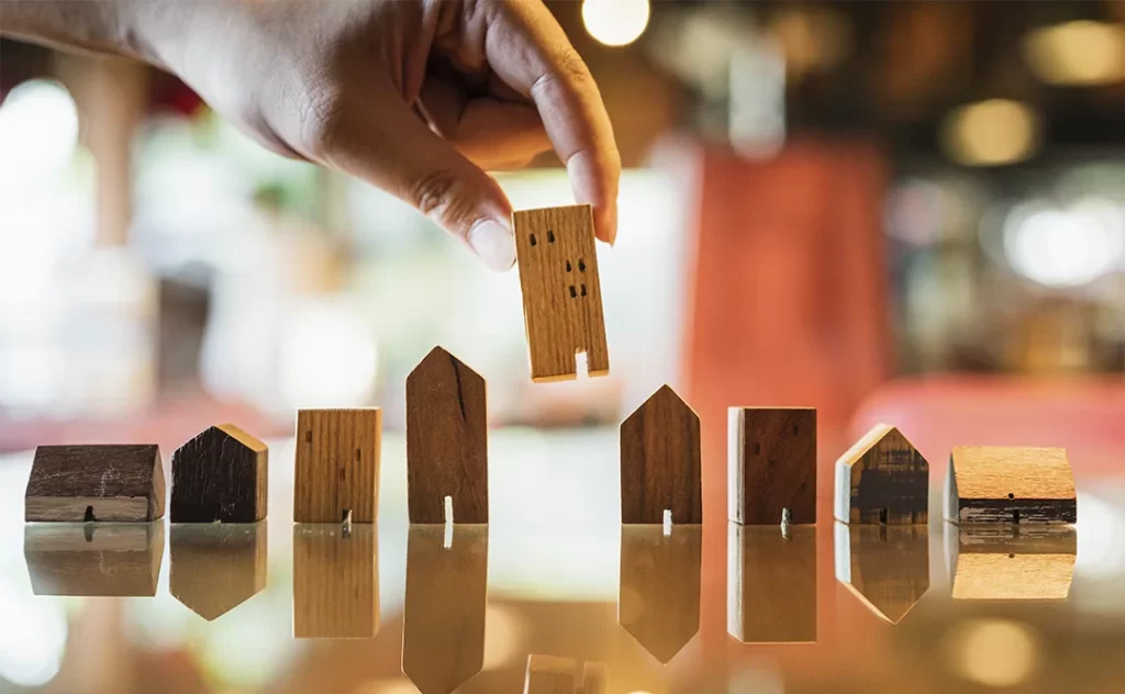 Indicadores imobiliários Indicadores imobiliários: A imagem mostra uma mão humana segurando um pequeno modelo de casa feito de madeira. Há outros modelos de casas de madeira de diferentes tamanhos e estilos dispostos em uma superfície, possivelmente um tábano ou uma superfície de madeira, com suas reflexões visíveis na superfície. A foto parece ter sido tirada em um ambiente interno com iluminação artificial, e a mão está na parte superior da imagem, destacando a interação com o modelo de casa.
