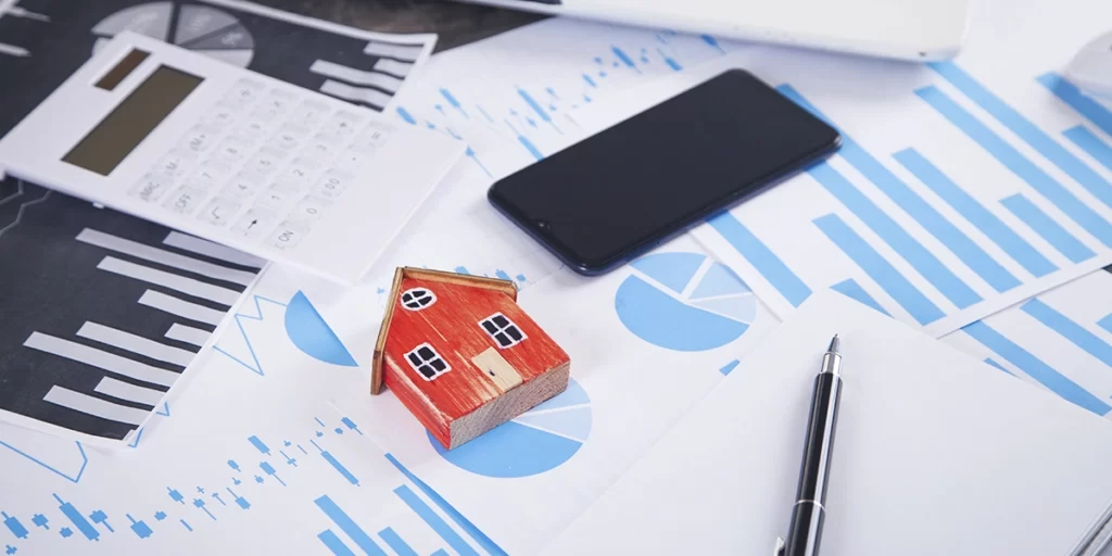 Índice Imobiliário Índice Imobiliário: A imagem mostra uma superfície de trabalho com vários itens relacionados a negócios e finanças. Há um aparelho de cálculo, um celular, um lápis, um pequeno modelo de casa, um caderno e algumas folhas de papel com gráficos e dados. A disposição dos itens sugere um ambiente de trabalho ou estudo financeiro ou de negócios.